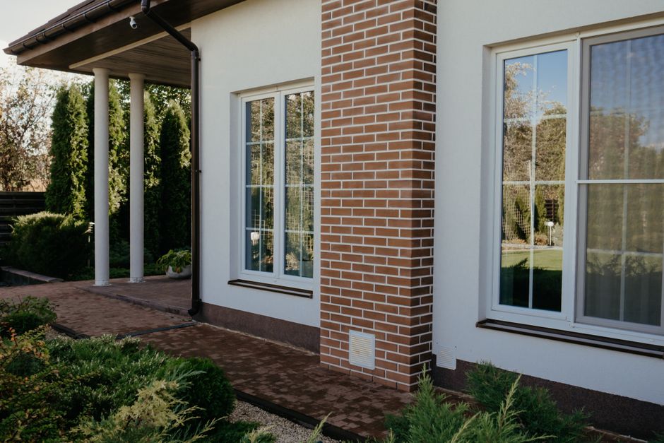 White painted house wall with two tall paned windows beside a brick column and low shrubs
