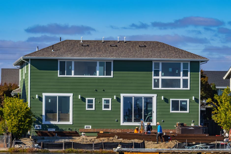 Back of a green sided two story house with several white framed windows and workers below