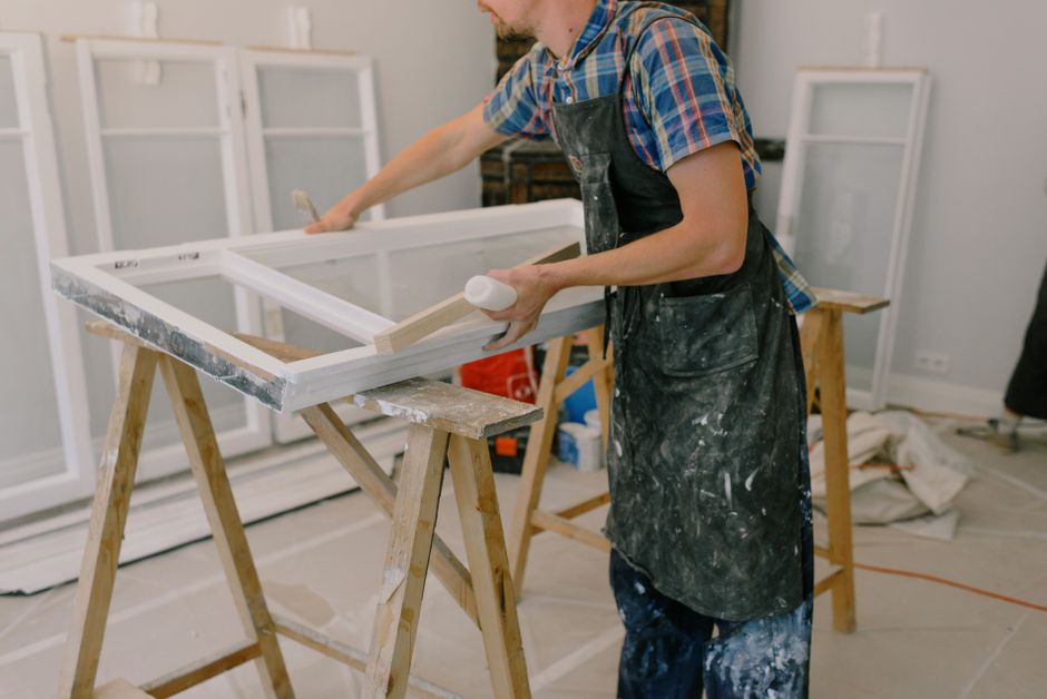 Worker in a paint spattered apron sanding a white window sash laid across sawhorses