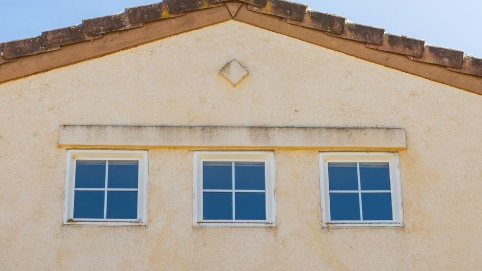 Three small white framed windows on a textured cream stucco gable end below a tile roof edge